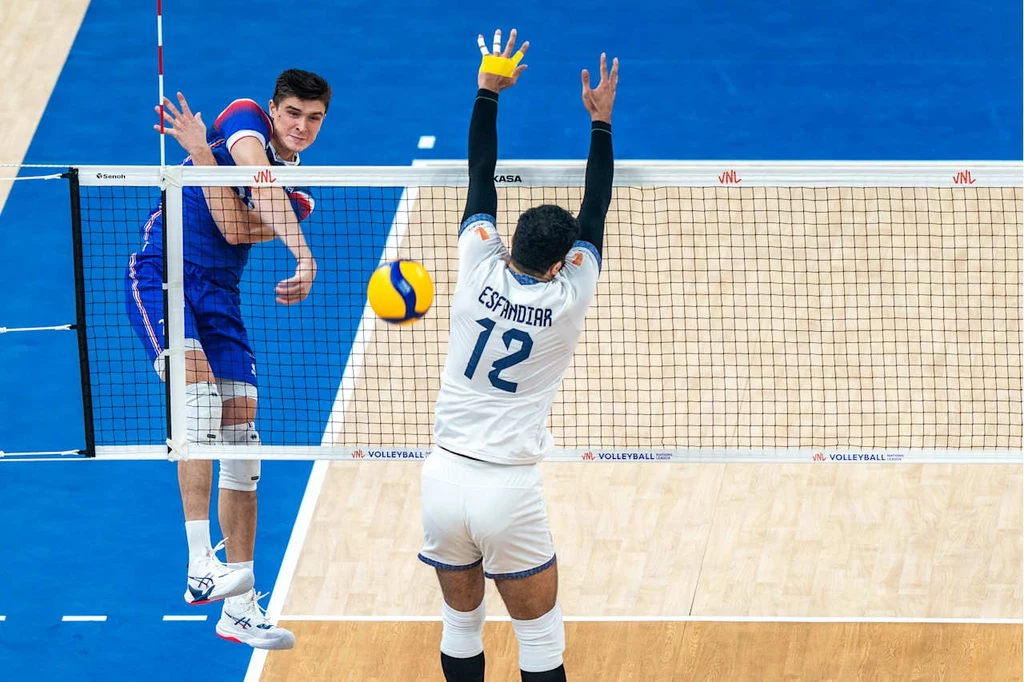 Liga Narodów siatkarzy. Kadr z meczu Francja - Iran Dwóch siatkarzy podczas dynamicznej akcji przy siatce, jeden z zawodników wykonuje atak, a drugi próbuje zablokować piłkę unosząc obie ręce nad siatką. Niebieskie boisko oraz piłka do siatkówki są wyraźnie widoczne.