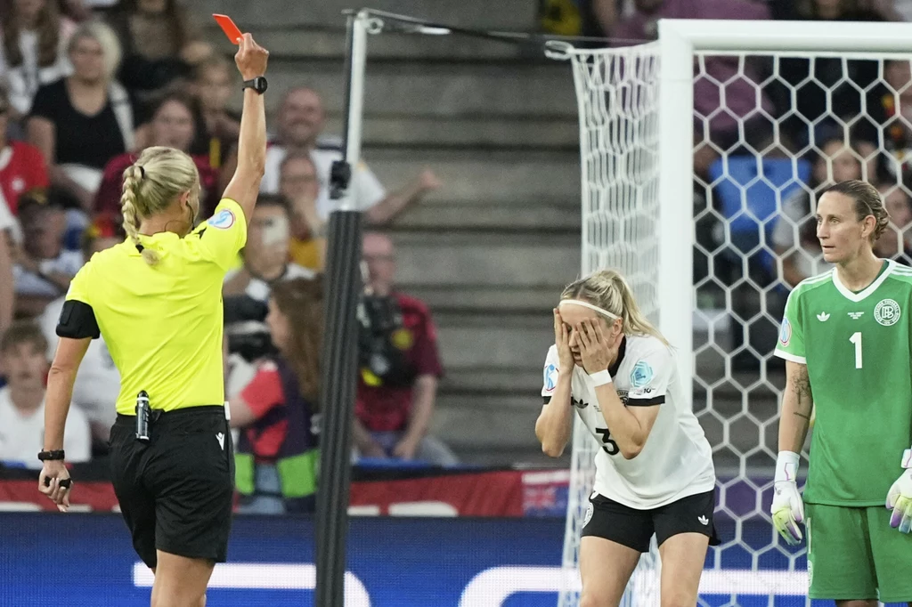 Kathrin Hendrich z czerwoną kartką w meczu z Francuzkami Sędzia pokazuje czerwoną kartkę zawodniczce drużyny w białych koszulkach, piłkarka zasłania twarz dłońmi w geście rozczarowania, obok stoi bramkarka w zielonym stroju przed bramką, w tle trybuny z widownią.