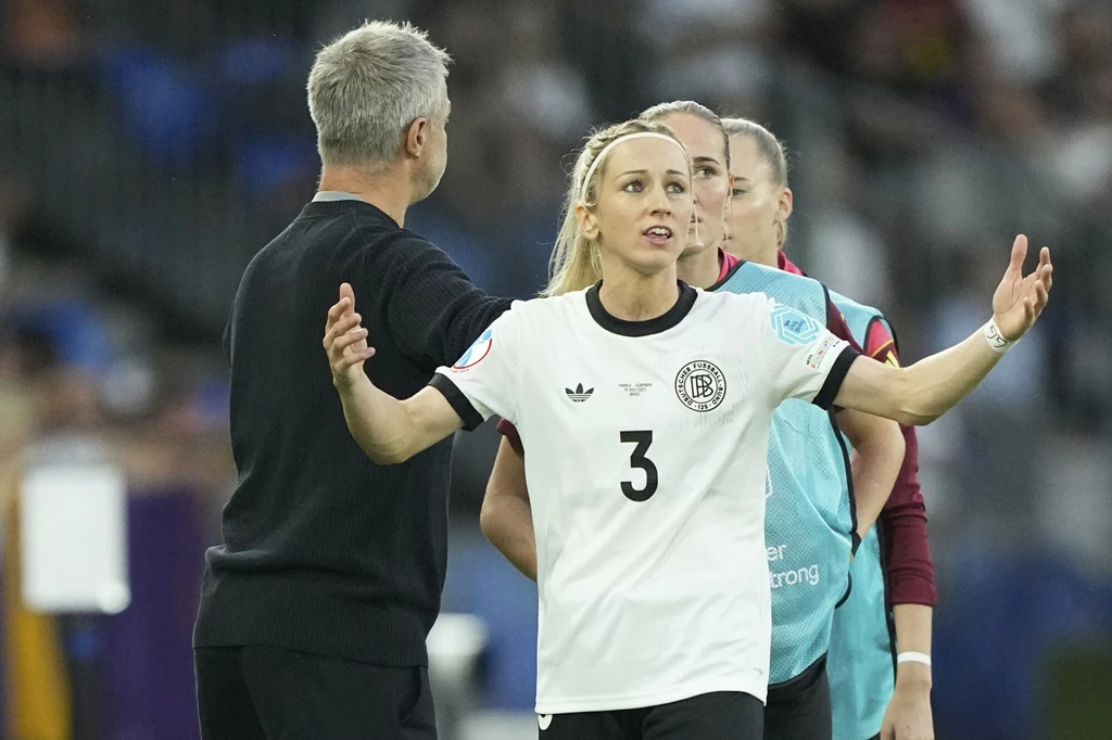 Kathrin Hendrich Zawodniczka w białej koszulce z numerem 3 i godłem reprezentacji Niemiec stoi z rozłożonymi rękami, obok niej trener oraz inne osoby z kadry, atmosfera wydaje się napięta jak podczas sportowej dyskusji.