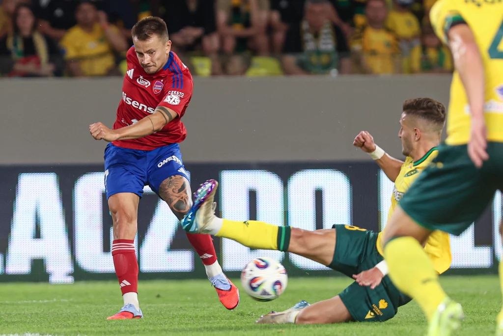 Raków - GKS Katowice Piłkarz drużyny w czerwonej koszulce uderza piłkę na bramkę, podczas gdy obrońca drużyny w zielono-żółtym stroju próbuje go zablokować wślizgiem, dynamiczna akcja rozgrywa się na tle trybun pełnych kibiców.