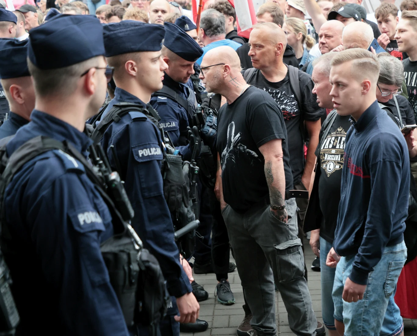 Policja podsumowała sobotnie antyimigranckie manifestacje zorganizowane przez Konfederację Tłum protestujących mężczyzn stoi naprzeciwko szpaleru uzbrojonych policjantów w mundurach, pośród których panuje napięta atmosfera, na twarzach uczestników widoczne emocje i determinacja.
