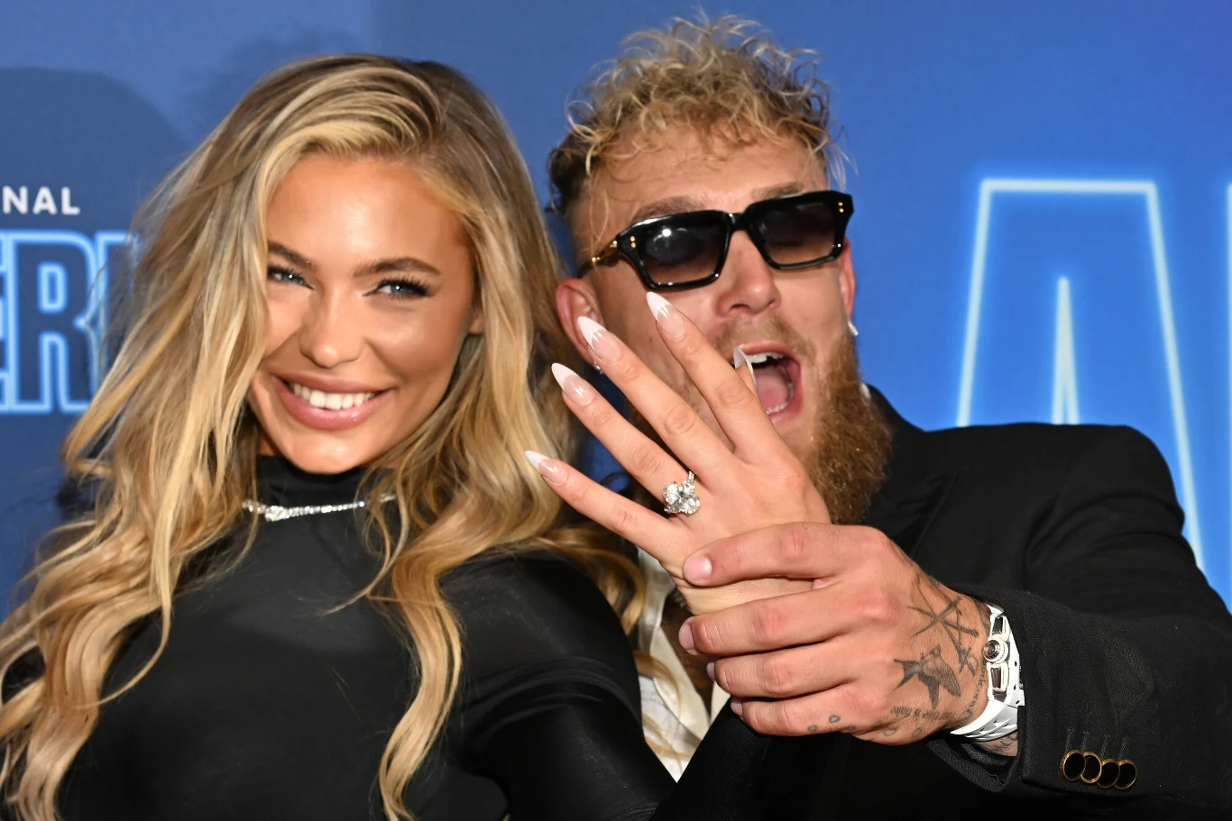 Jutta Leerdam i Jake Paul Para uśmiecha się szeroko, mężczyzna pokazuje dłonią pierścionek zaręczynowy założony na palec kobiety, oboje ubrani elegancko, na niebieskim tle z elementami napisu.