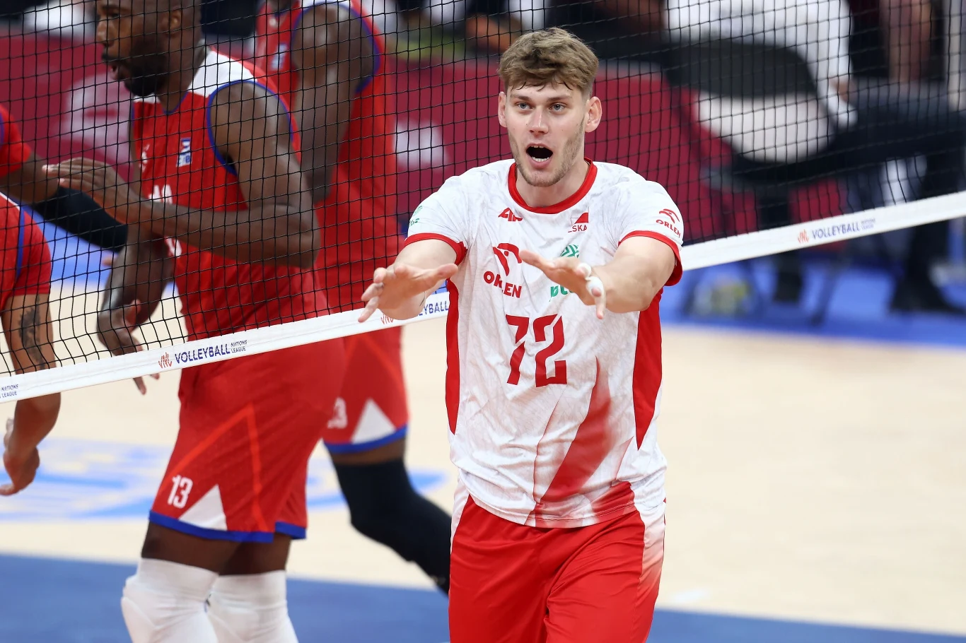 Liga Narodów siatkarzy. Mateusz Poręba zasłużył na pochwałę od Grbicia Siatkarz w białoczerwonym stroju z numerem 72 wykonuje ruch przy siatce podczas meczu, w tle widoczni zawodnicy drużyny przeciwnej w niebiesko-czerwonych strojach.