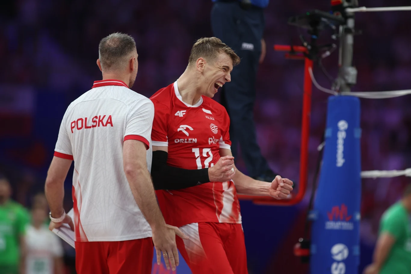 Siatkarz w czerwonym stroju energetycznie świętuje zdobycie punktu podczas meczu, obok stoi trener reprezentacji Polski w białoczerwonej koszulce, w tle widoczne elementy boiska siatkarskiego.