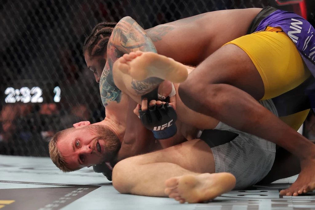 UFC: Łukasz Brzeski - Ryan Spann Dwóch zawodników MMA walczy na macie w oktagonie, jeden z nich próbuje zastosować technikę grapplingu, przyciskając przeciwnika do ziemi, wokół widać siatkę zabezpieczającą oraz inne elementy areny