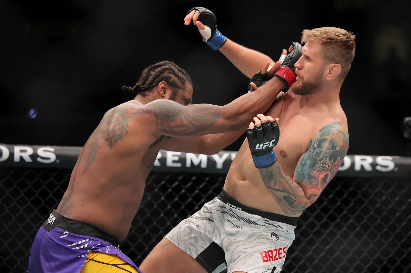 UFC: Ryan Spann - Łukasz Brzeski Dwóch zawodników UFC walczy w oktagonie, jeden z nich wyprowadza cios prosto w twarz przeciwnika. Obaj są umięśnieni i skupieni na rywalizacji, jeden z zawodników ma wyraźne tatuaże na ramieniu i klatce piersiowej.