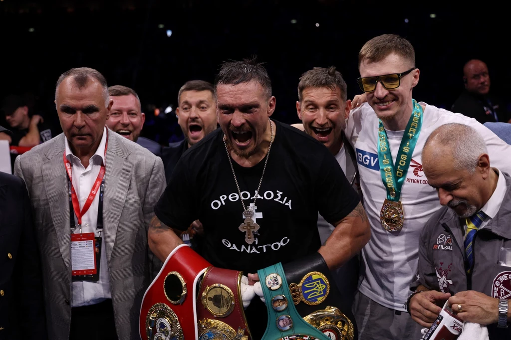 Ołeksandr Usyk Grupa mężczyzn świętuje zwycięstwo boksera na ringu, centralnie znajduje się sportowiec ubrany w czarną koszulkę i z kilkoma pasami mistrzowskimi przewieszonymi przez ramiona, wokół stoją członkowie ekipy oraz kibice z medalami.