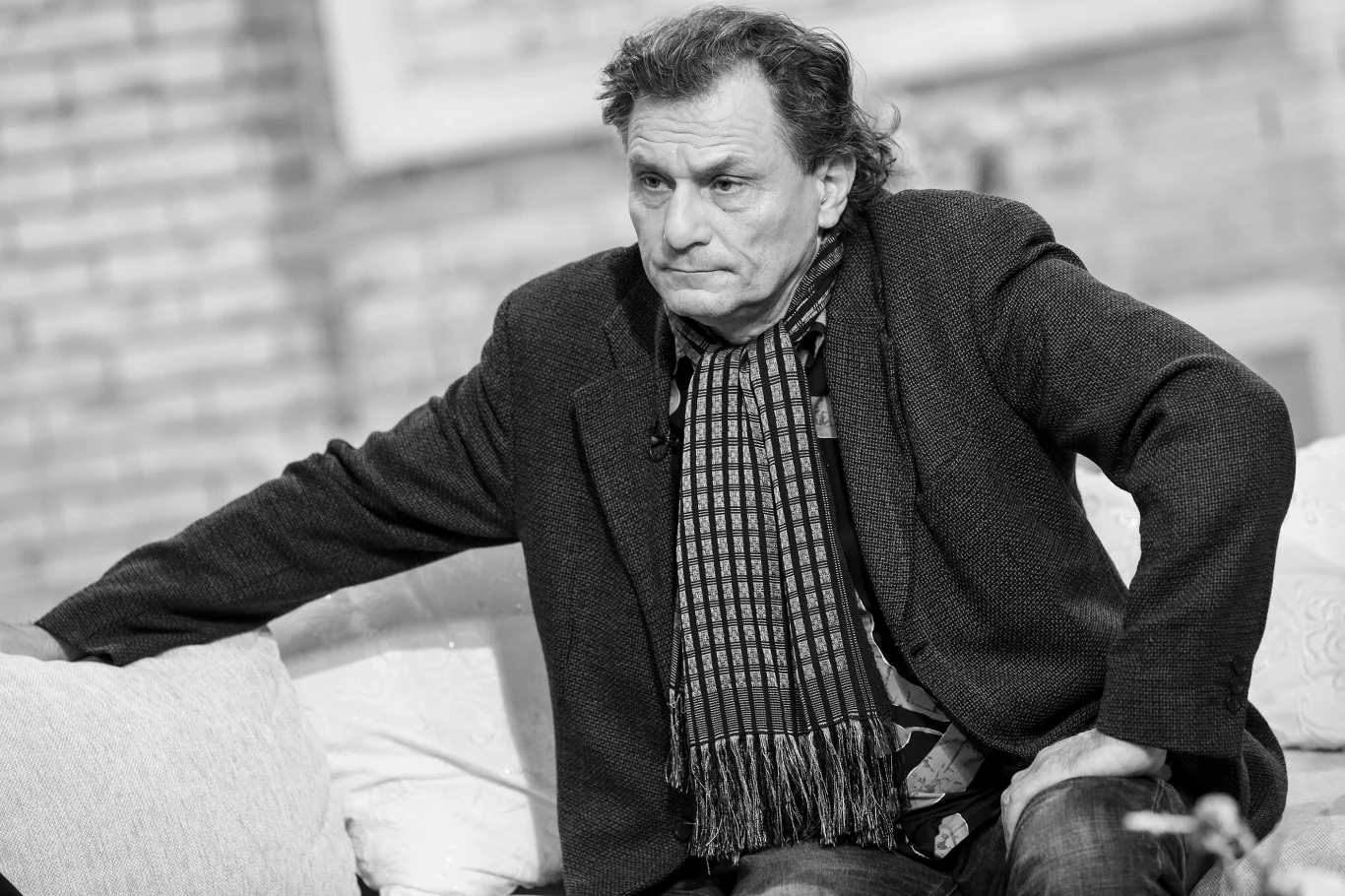 Ryszard Olesiński (exMaanam) miał 71 lat Mężczyzna w średnim wieku z lekko posiwiałymi włosami siedzi na kanapie, ubrany w płaszcz i szalik w kratę, z poważnym wyrazem twarzy, jedno ramię opiera o poręcz sofy.