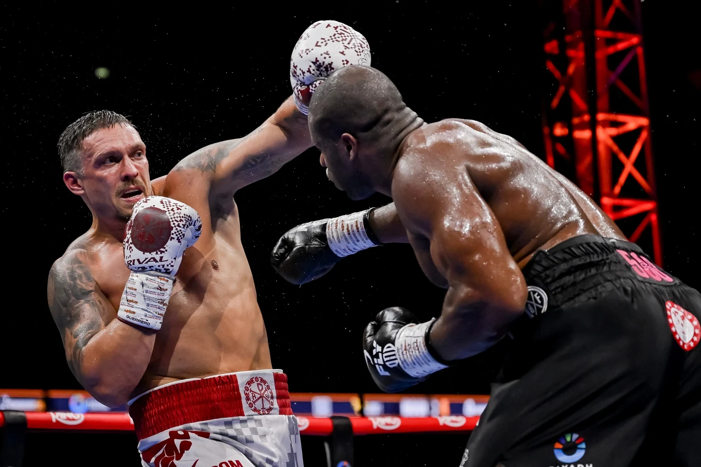 Ołeksandr Usyk i Daniel Dubois Dwóch bokserów w trakcie pojedynku w ringu, jeden z nich unika ciosu wyprowadzanego przez rywala, wokół panuje sportowa atmosfera, widoczne światła areny sportowej.