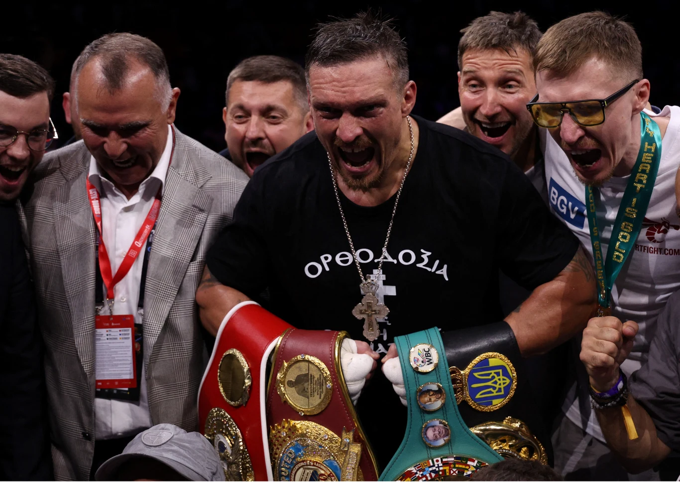 Grupa mężczyzn wyraża silne emocje podczas świętowania zwycięstwa, centralna postać w czarnej koszulce i z medalami oraz pasami mistrzowskimi w rękach stoi na pierwszym planie, wyraźnie pokazując radość i triumf.