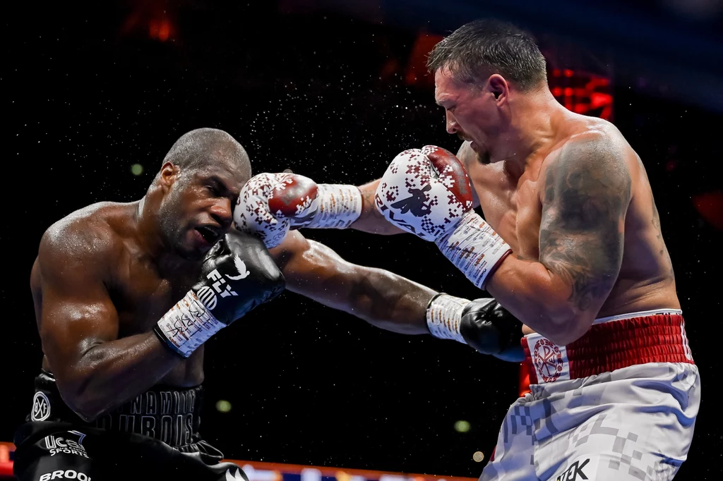 Ołeksandr Usyk był bezlitosny dla Daniela Duboisa Mocny cios zadany przez boksera w czerwonych rękawicach trafia przeciwnika w twarz, dynamiczna scena na ringu bokserskim, zawodnicy w sportowych strojach, wyraźnie widoczne emocje oraz wysiłek fizyczny.