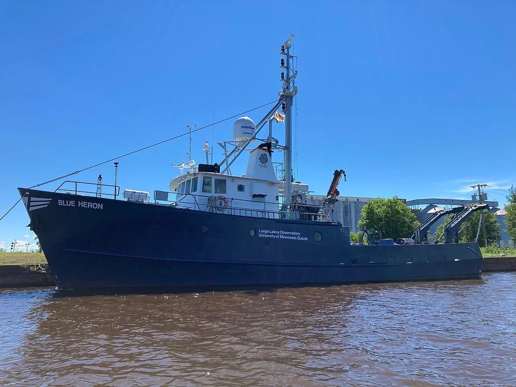 Statek "Blue Heron" bada życie Wielkich Jezior Amerykańskich Statek badawczy o nazwie BLUE HERON zacumowany przy brzegu, wyróżniający się nowoczesnym osprzętem na pokładzie, masztami oraz widocznymi urządzeniami satelitarnymi; w tle widać drzewa i fragmenty miejskiej zabudowy.