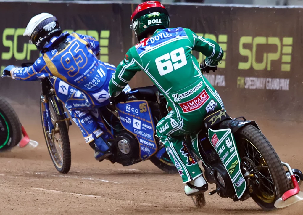 Dwóch zawodników rywalizujących na torze żużlowym podczas dynamicznego momentu wyścigu, ubranych w kolorowe kombinezony i kaski ochronne, pochylonych na motocyklach w trakcie pokonywania zakrętu.