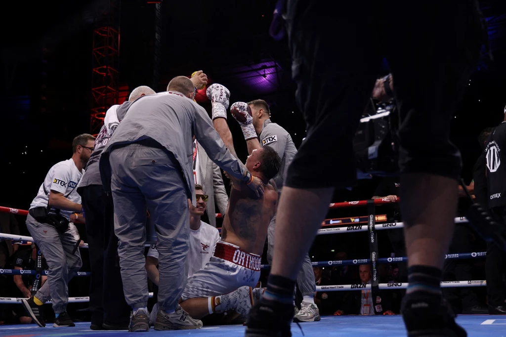 Ołeksandr Usyk po walce z Danielem Dubois Bokser klęczący na ringu otoczony przez kilku członków ekipy technicznej, jeden z nich unosi jego rękę w górę. Widoczne liny ringu i światła na arenie, całość w dynamicznej sportowej atmosferze.