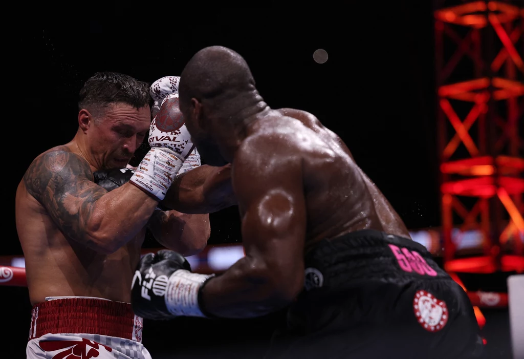 Walka Ołeksandr Usyk vs Daniel Dubois Dwóch bokserów walczy ze sobą na ringu, jeden z nich zadaje silny cios przeciwnikowi, który osłania twarz rękoma, tło stanowią elementy oświetlenia i infrastruktury hali sportowej.