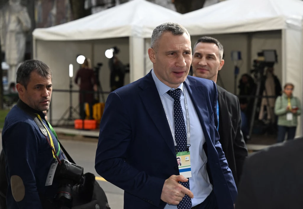 Witalij Kliczko Mężczyzna w garniturze z identyfikatorem na szyi idący ulicą, w tle białe namioty oraz osoby z aparatami fotograficznymi i kamerami, sugerujące obecność mediów.