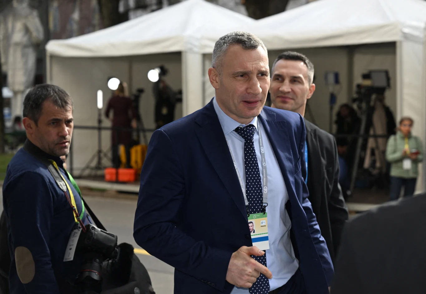 Mężczyzna w garniturze z identyfikatorem na szyi idący ulicą, w tle białe namioty oraz osoby z aparatami fotograficznymi i kamerami, sugerujące obecność mediów.