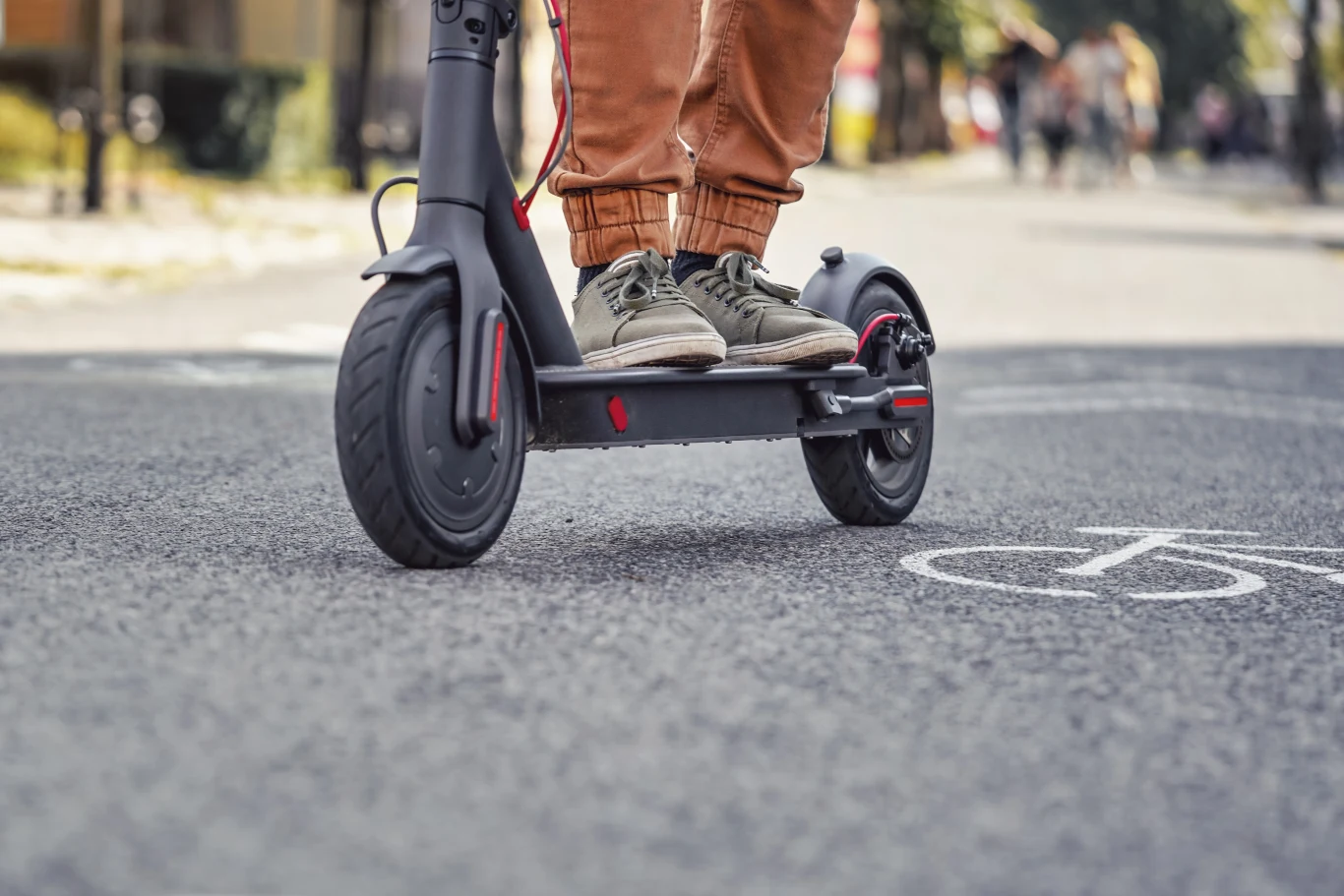 16-latek bez uprawnień potrącił elektryczną hulajnogą rowerzystkę (fot. ilustracyjne) Osoba w sportowych butach i brązowych spodniach stoi na elektrycznej hulajnodze jadącej po asfaltowej drodze z oznaczeniem ścieżki rowerowej.