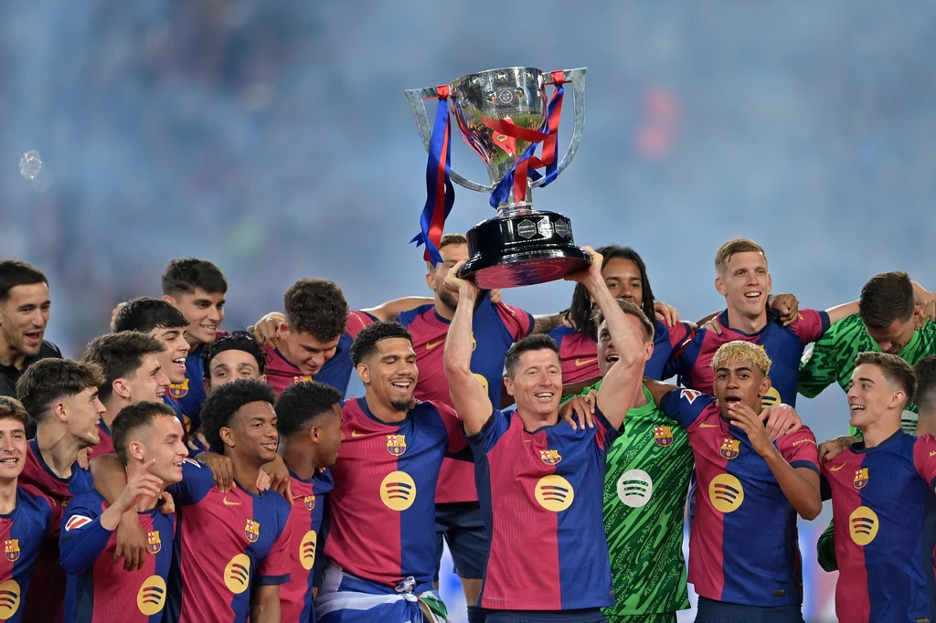 Piłkarze Barcelony z trofeum za mistrzostwo Hiszpanii Piłkarze drużyny FC Barcelona ubrani w klubowe stroje świętują zwycięstwo, jeden z zawodników unosi w górę dużą srebrną trofeum owiniętą w klubowe wstążki, wszyscy są uśmiechnięci i radośni, w tle rozmyta niebieska scenografia.