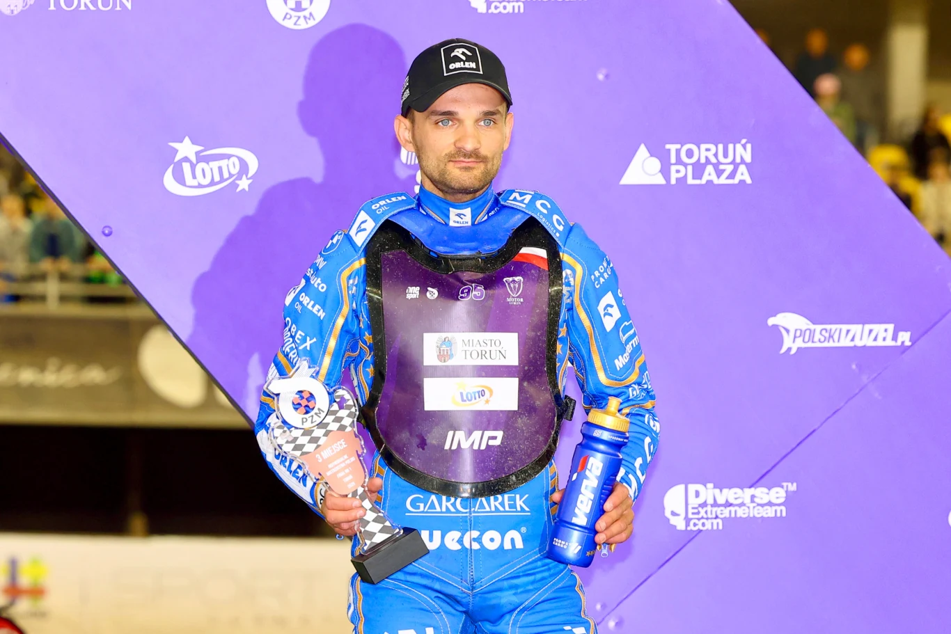 Zawodnik sportów motocyklowych ubrany w niebieski kombinezon wyścigowy stoi na tle fioletowej ścianki z logotypami sponsorów, w jednej ręce trzyma puchar, w drugiej bidon, wokół widoczne jasne światła i elementy scenografii zawodów