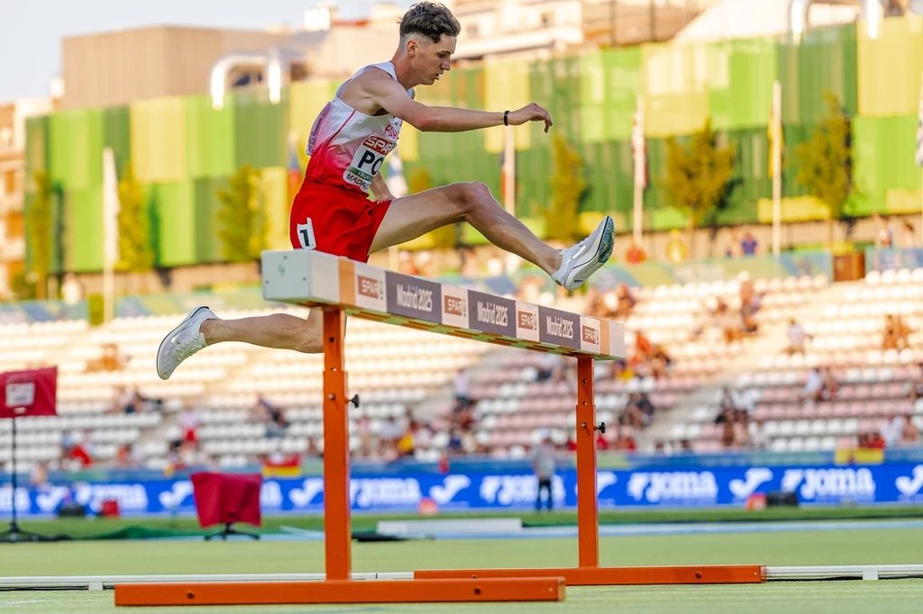 Lekkoatleta w biało-czerwonym stroju podczas biegu z przeszkodami przeskakuje płotek na stadionie lekkoatletycznym, w tle trybuny z widzami oraz miejskie budynki.