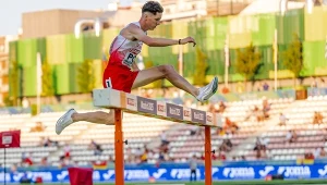 Lekkoatleta w biało-czerwonym stroju podczas biegu z przeszkodami przeskakuje płotek na stadionie lekkoatletycznym, w tle trybuny z widzami oraz miejskie budynki.