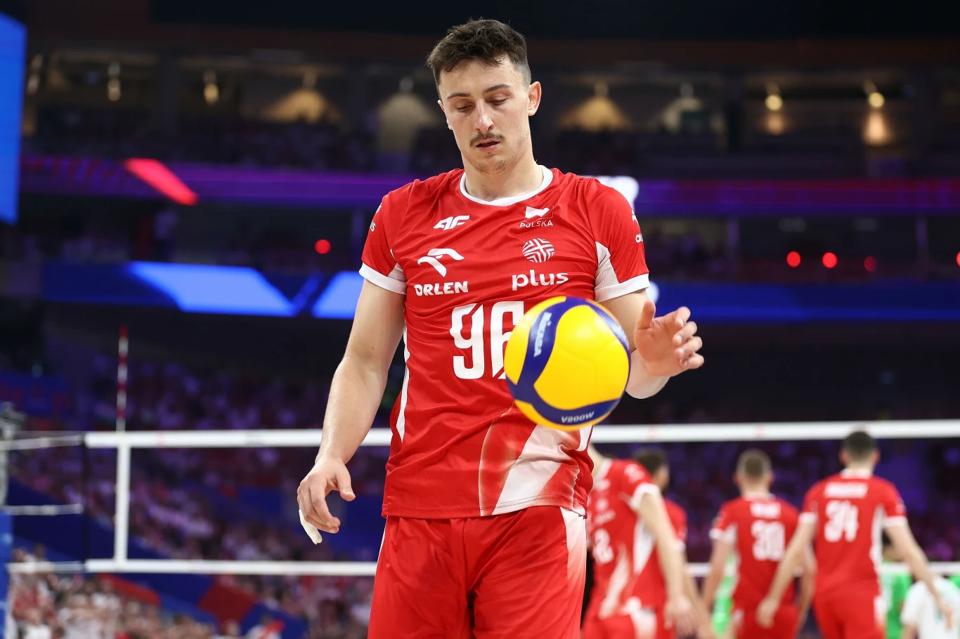 Jan Firlej Zawodnik polskiej reprezentacji siatkówki w czerwonym stroju przygotowuje się do serwisu podczas meczu na hali, w tle widoczni są inni siatkarze oraz siatka.