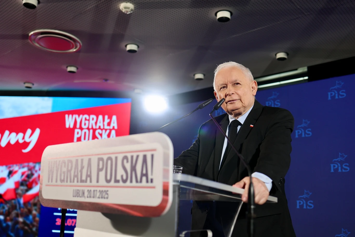 Jarosław Kaczyński zapowiedział prace nad nowym programem wyborczym PiS Starszy mężczyzna w garniturze stojący przy mównicy podczas konferencji prasowej, w tle widoczna grafika z napisem 'WYGRAŁA POLSKA' oraz logo partii politycznej, scena oświetlona profesjonalnym światłem.