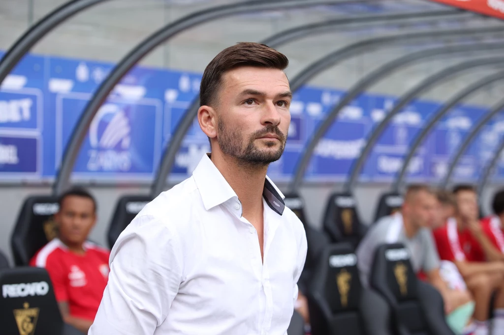 W niedzielne popołudnie Michal Gasparik zadebiutował w roli pierwszego szkoleniowca Górnika Zabrze Mężczyzna w białej koszuli stoi na tle stadionowej ławki rezerwowych, w tle kilku piłkarzy siedzi na ławkach w czerwonych strojach sportowych.