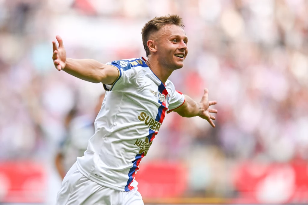 Kamil Lukoszek na liście strzelców. W tym momencie gospodarze prowadzili 1:0 Piłkarz w białym stroju z rozłożonymi ramionami świętuje zdobycie gola na tle rozmytego stadionu pełnego kibiców.