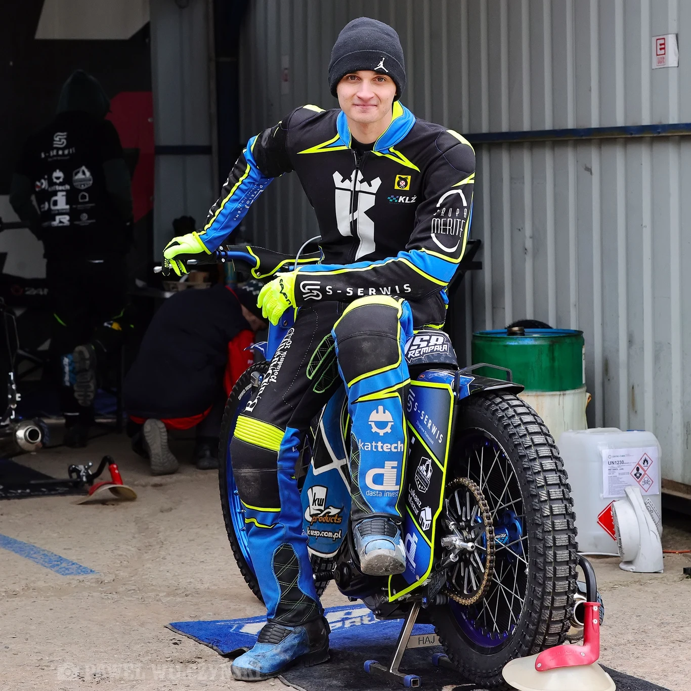 Zawodnik sportów motorowych w kolorowym kombinezonie, siedzący na motocyklu żużlowym w warsztacie lub garażu, w tle widoczne są narzędzia, pojemniki i inne osoby związane z obsługą motocykli.