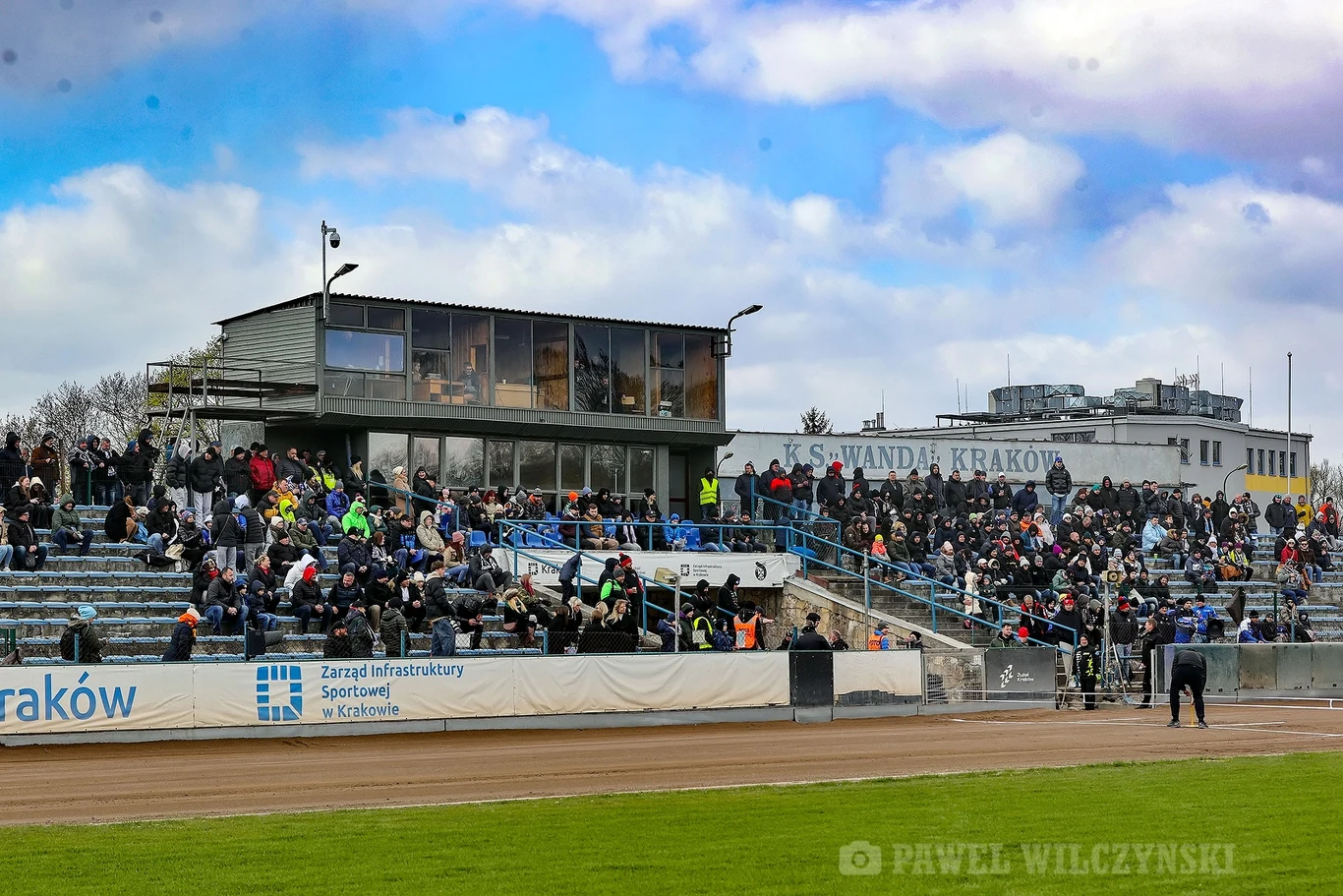 Tłum ludzi zgromadzony na trybunach stadionu sportowego, na pierwszym planie zadaszony budynek z przeszklonymi oknami, a w tle widoczny napis 'Wanda Kraków'.