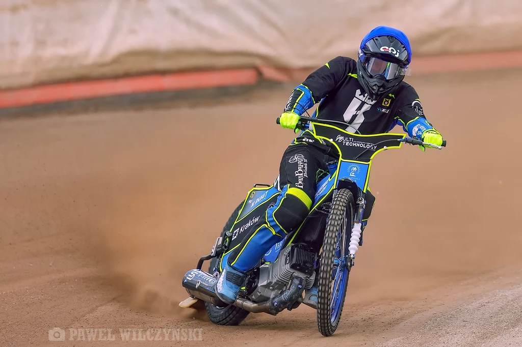 Zawodnik sportów motorowych w pełnym ochraniaczu i kasku prowadzi motocykl żużlowy na torze, dynamicznie pokonując zakręt, podkreślając sportowy charakter sytuacji.