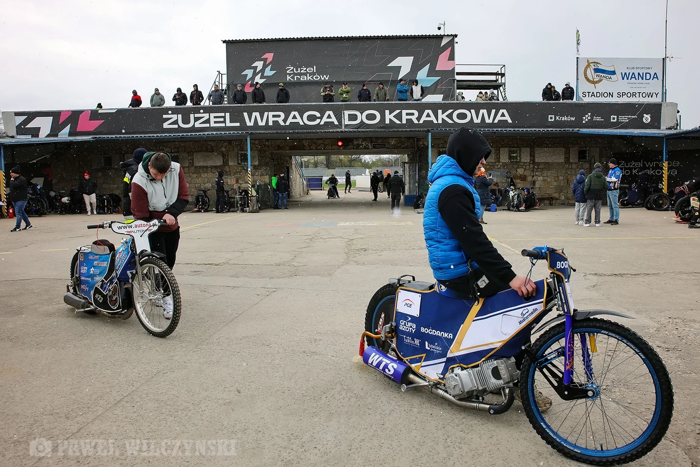 Dwóch zawodników na motocyklach żużlowych przygotowuje się na stadionie żużlowym w Krakowie, w tle widać trybuny oraz baner z napisem 'Żużel wraca do Krakowa', za motocyklistami kibice i obsługa stadionu.