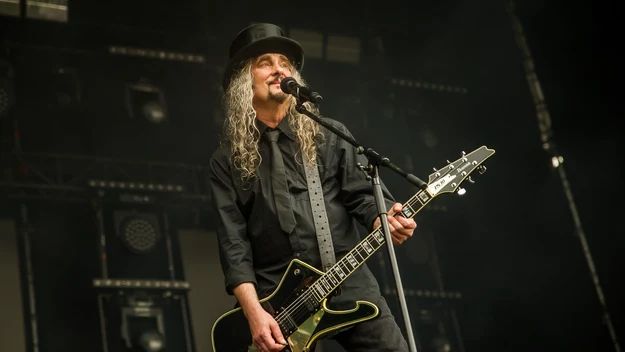 Mężczyzna z długimi, siwymi włosami i w czarnym kapeluszu gra na gitarze elektrycznej i śpiewa do mikrofonu na scenie koncertowej, ubrany na czarno, w tle oświetlenie sceniczne.