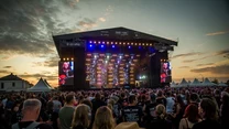 Tłum zgromadzony przed dużą sceną koncertową podczas plenerowego festiwalu muzycznego o zmierzchu, scena oświetlona kolorowymi światłami, wyświetlacze LED z wizerunkiem muzyków i dynamiczne niebo w tle.