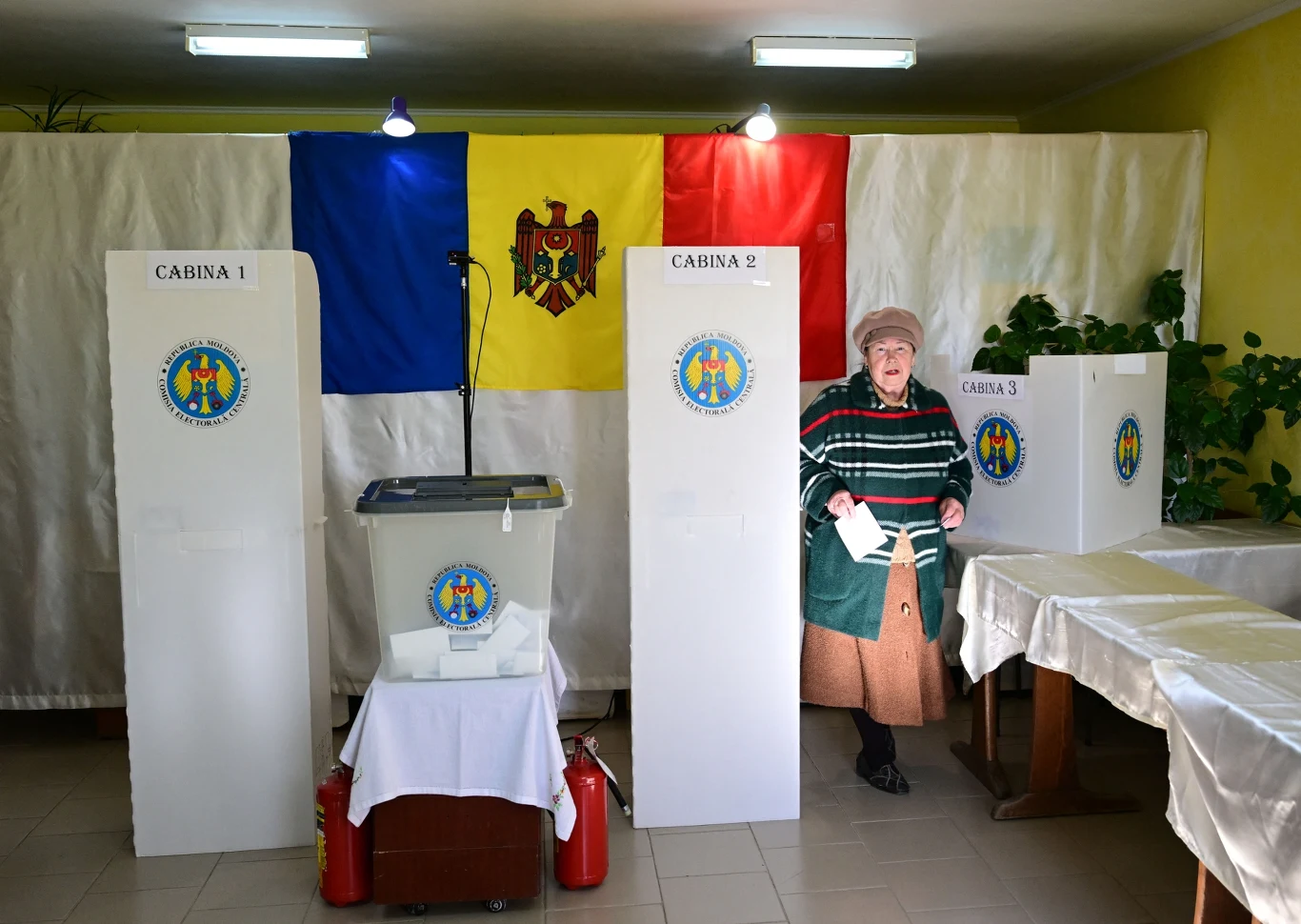 Prorosyjska koalicja zablokowana przez CKW Starsza kobieta opuszcza kabinę do głosowania, trzymając kartę wyborczą w ręku. Tło stanowi rozwieszona flaga Mołdawii, po lewej stronie widoczna urna wyborcza oraz trzy oddzielone kabiny. W pomieszczeniu panuje jasne oświetlenie, a obok znajdują się r...