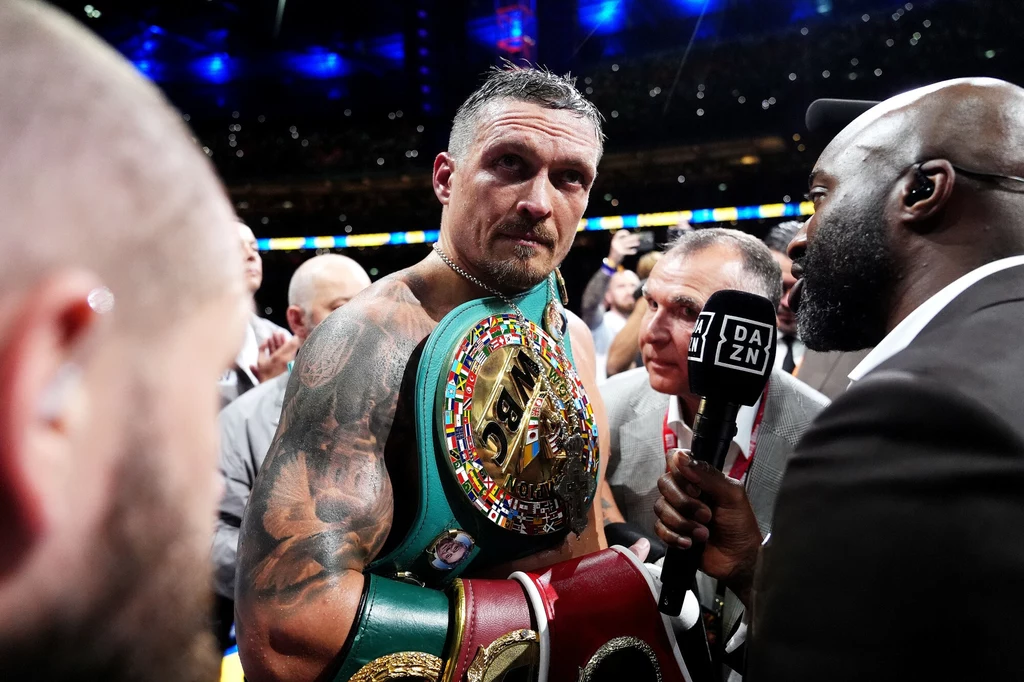 Oleksandr Usyk Bokser z odsłoniętym torsem trzyma na ramieniu kilka mistrzowskich pasów bokserskich, wokół niego zgromadzeni są dziennikarze z mikrofonami, za nim widoczna publiczność na stadionie.