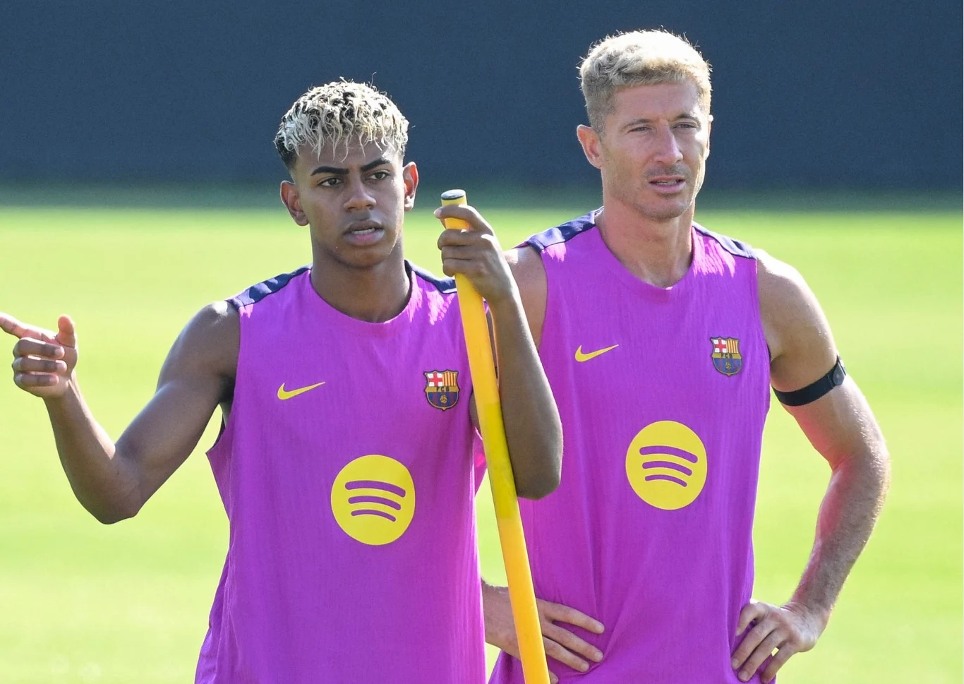 Lamine Yamal i Robert Lewandowski Dwóch piłkarzy FC Barcelony w różowo-fioletowych strojach treningowych stoi na boisku, jeden opiera się o żółty drążek, drugi wskazuje ręką, w tle rozmyta murawa.