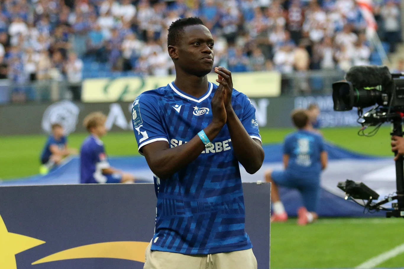 Timothy Ouma, nowy pomocnik Lecha Poznań Piłkarz w niebieskiej koszulce drużyny Lech Poznań stoi na boisku z rękami złożonymi w geście podziękowania, w tle rozmazana publiczność na trybunach i inni zawodnicy.