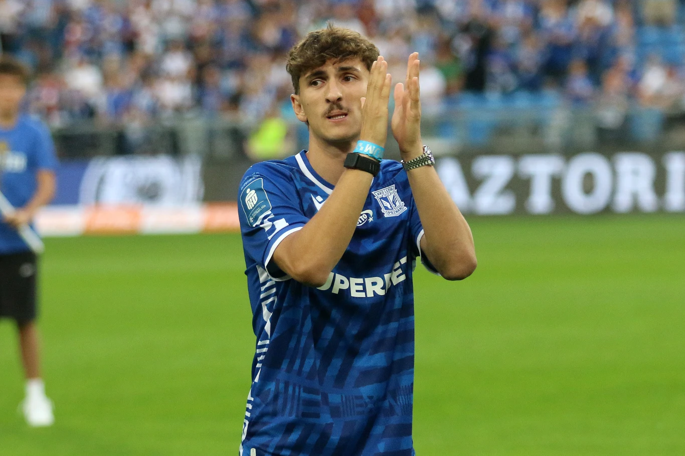 Pablo Rodriguez, nowy zawodnik Lecha Poznań Młody piłkarz ubrany w niebieską koszulkę drużyny Lech Poznań znajduje się na murawie stadionu, klaszcząc w dłonie, w tle widownia i inni zawodnicy rozmazani ze względu na głębię ostrości.