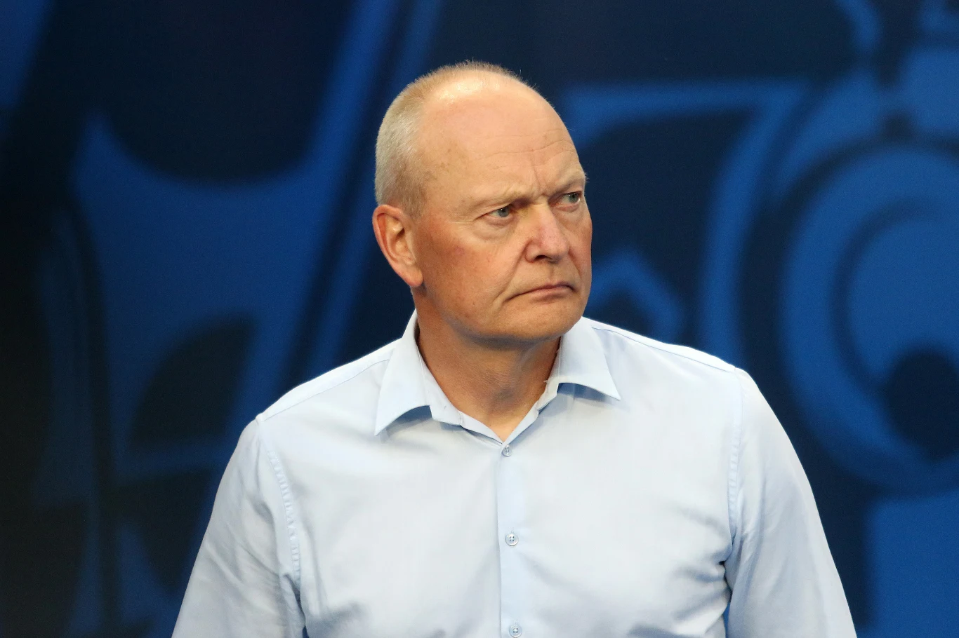 Niels Frederiksen, trener Lecha Poznań Mężczyzna w średnim wieku ubrany w jasnoniebieską koszulę patrzy z powagą w bok na tle ciemnego, abstrakcyjnego wzoru.