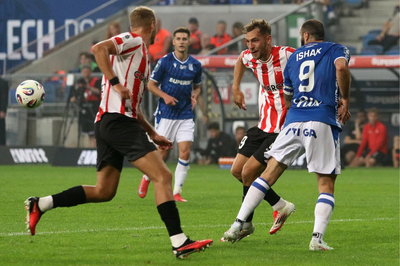 Dynamiczna akcja podczas meczu piłki nożnej, zawodnik w niebieskiej koszulce próbuje kopnąć piłkę, zawodnicy w biało-czerwonych strojach próbują go zablokować, w tle widoczna publiczność i fragment stadionu.