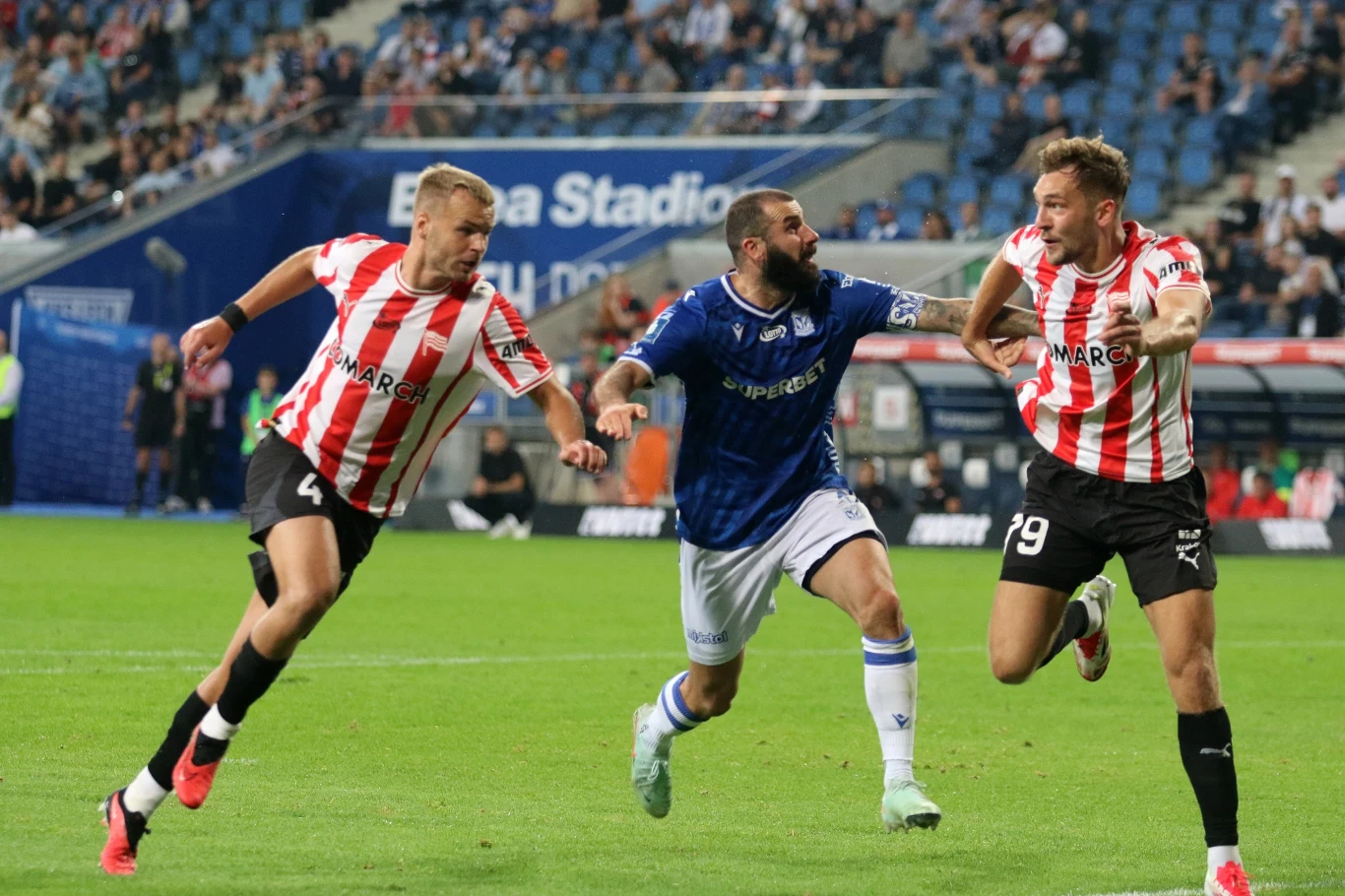 Trzech piłkarzy walczy o piłkę na murawie podczas meczu piłkarskiego, dwóch z nich w biało-czerwonych koszulkach ściga się z zawodnikiem drużyny przeciwnej w niebieskim stroju; w tle kibice i fragment stadionu.