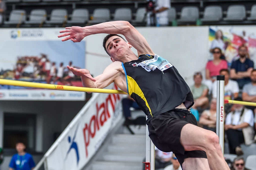 Lekkoatleta w czarnej koszulce z żółtymi akcentami pokonuje poprzeczkę podczas skoku wzwyż na stadionie, w tle widownia oraz banery reklamowe.