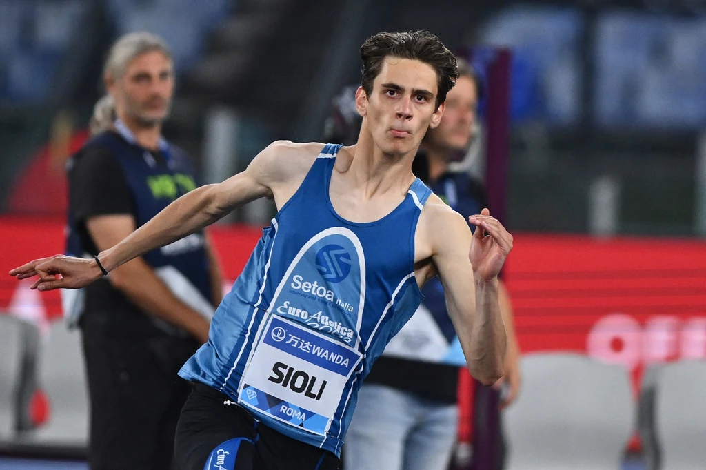 Młody sportowiec w niebieskim stroju lekkoatletycznym w trakcie rzutu, skupiony wyraz twarzy, na torsie widoczny numer i nazwisko, w tle inni uczestnicy zawodów lekkoatletycznych oraz rozmyte trybuny stadionu.