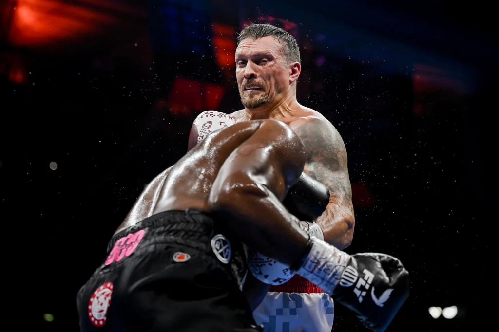 Ołeksandr Usyk Dwóch bokserów w trakcie walki na ringu, jeden z nich zadaje mocny cios przeciwnikowi w korpus, na tle ciemnej hali sportowej i świateł.
