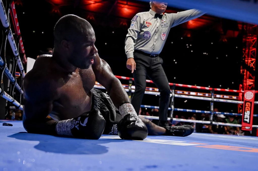 Ołeksandr Usyk Bokser siedzi na macie ringu, ewidentnie po silnym ciosie, w tle sędzia odlicza czas i obserwuje sytuację, wokół widoczne elementy ringu i światła areny.