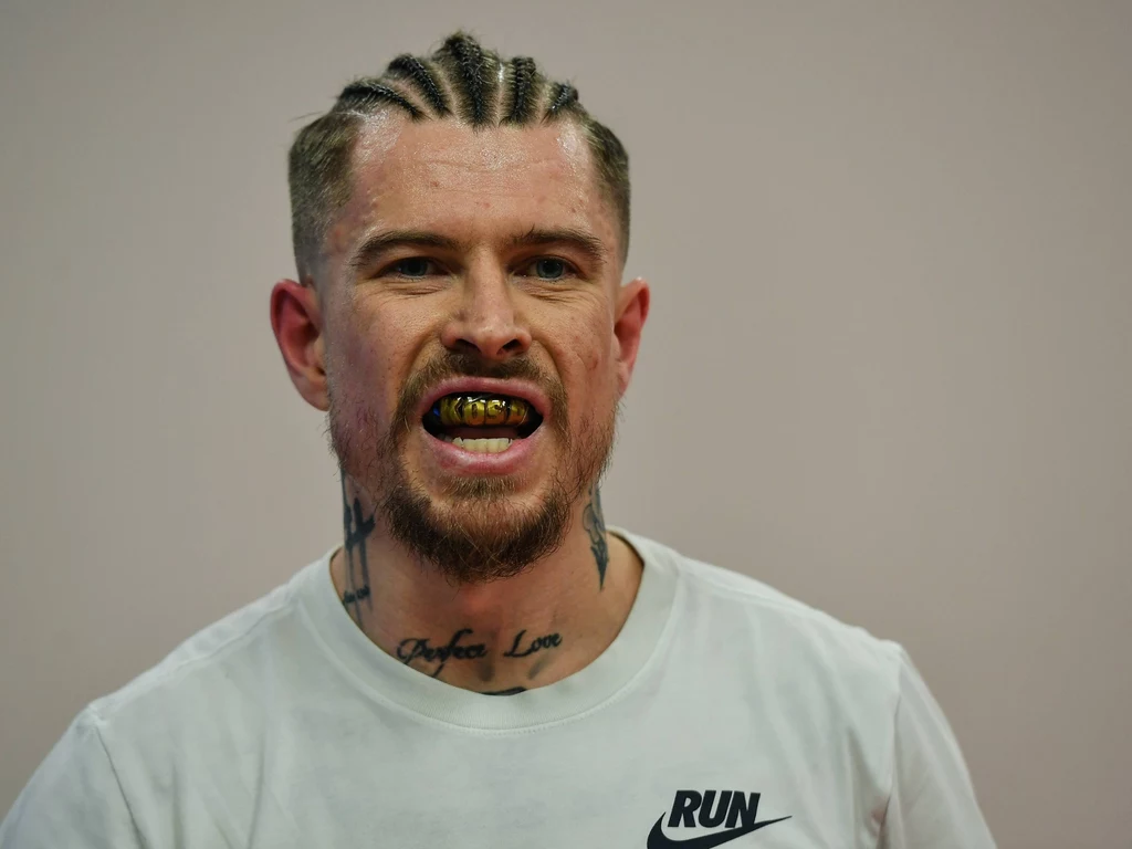 Jakub Kosecki Mężczyzna z dredami na głowie, licznymi tatuażami na szyi i uzębieniem z grillami w kolorze złotym, ubrany w białą koszulkę sportową.