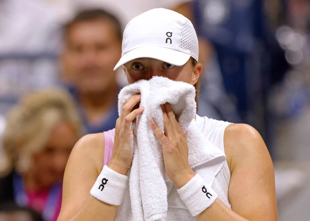 Iga Świątek już przygotowuje się do tegorocznej edycji US Open Tenisistka w białej czapce i stroju sportowym wyciera twarz ręcznikiem na korcie, widoczne skupienie podczas przerwy w meczu.